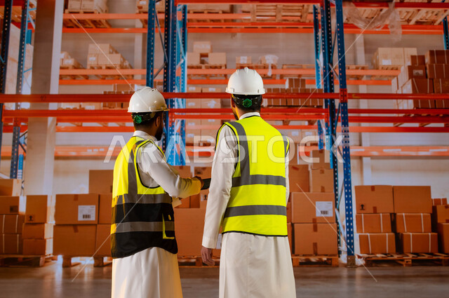 استخدام التقنية والتكنولوجيا لتسجيل الحسابات وجرد البضائع في المستودع، بضائع ومنتجات عربية سعودية خليجية, تصوير من الخلف لمدير ترتيب وتنظيم المستودعات المحلية ومهندس عربي سعودي خليجي، خلفية صناديق وكراتين مغلفة.