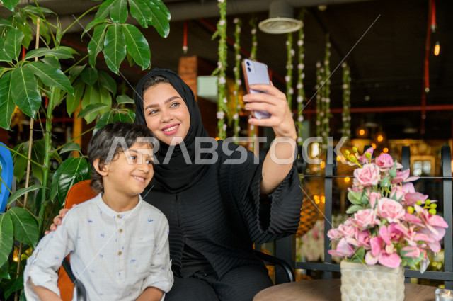 طفل عربي سعودي خليجي يقضي وقته مع امه في المطعم بايماءات و تعابير وجه مختلفة ، تلتلقط الام صور سلفي لها و لأبنها بكاميرا هاتفها المحمول، صورة تذكارية ، مطعم سعودي 