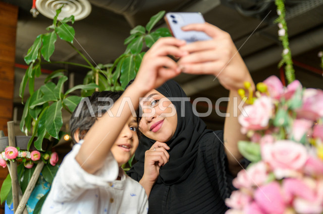 طفل عربي سعودي خليجي يقضي وقته مع امه في المطعم بايماءات و تعابير وجه مختلفة ، تلتلقط الام صور سلفي لها و لأبنها بكاميرا هاتفها المحمول، صورة تذكارية ، مطعم سعودي 