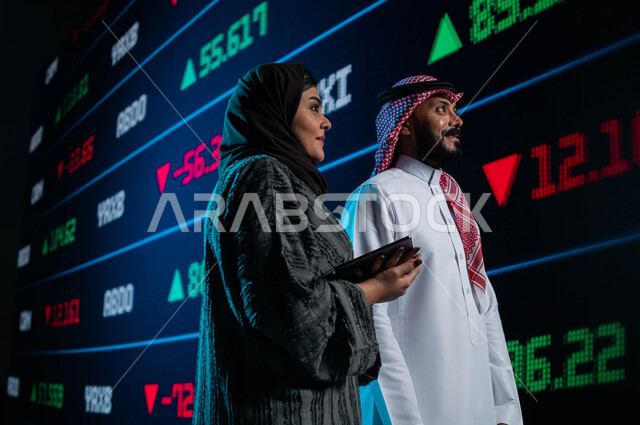 Follow-up of the rise and fall of stocks through modern electronic screens, the use of technology to follow the stock market, a Saudi Arabian Gulf businessman and businesswoman discussing the stock market and using a portable tablet, financial and economi