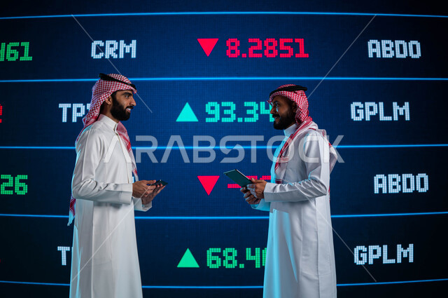 Discussion and follow-up of digital data and information for Saudi stocks, two Saudi Gulf Arab businessmen using a tablet, financial and economic investment in the Saudi Stock Exchange, follow-up on the stock market, a modern electronic screen showing the