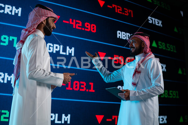 الإستثمار المالي والإقتصادي في البورصة السعودية، رجلان أعمال سعوديان خليجيان عربيان يستخدمان التابليت ويتناقشان في البيانات والمعلومات الرقمية للأسهم السعودية، متابعة سوق الاوراق المالية، شاشة إلكترونية حديثة تظهر الأسهم المتداول بها في السعودية