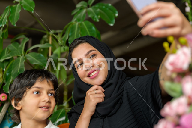 طفل عربي سعودي خليجي يقضي وقته مع امه في المطعم بايماءات و تعابير وجه مختلفة ، تلتلقط الام صور سلفي لها و لأبنها بكاميرا هاتفها المحمول، صورة تذكارية ، مطعم سعودي 
