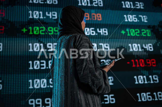 متابعة سوق الاوراق المالية من خلال شاشات الكترونية حديثة، سيدة أعمال عربية خليجية سعودية ترتدي نظارات الواقع الافتراضي وتقف امام شاشة البورصة، إستخدام التابليت في جمع البيانات والمعلومات الرقمية للأسهم، الإستثمار المالي والإقتصادي في البورصة السعودية