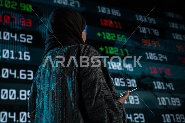 متابعة الأسهم المتداول بها في السعودية من خلال شاشات الكترونية حديثة، سيدة أعمال عربية خليجية سعودية ترتدي نظارات الواقع الافتراضي وتقف امام شاشة البورصة، إستخدام التابليت في جمع البيانات والمعلومات الرقمية، الإستثمار المالي والإقتصادي في البورصة السعودية
