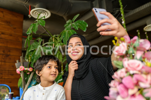 طفل عربي سعودي خليجي يقضي وقته مع امه في المطعم بايماءات و تعابير وجه مختلفة ، تلتلقط الام صور سلفي لها و لأبنها بكاميرا هاتفها المحمول، صورة تذكارية ، مطعم سعودي 