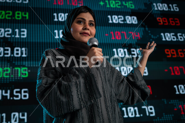 عرض تقديمي لأخبار أسهم الشركات في البورصة السعودية، سيدة اعمال خليجية سعودية عربية تمسك المايكروفون وتتحدث في مؤتمر اقتصادي، ندوات اقتصادية سعودية، مقترحات ونصائح حول التدوال الإلكتروني، خلفية شاشات الكترونية حديثة لمتابعة السوق السعودي