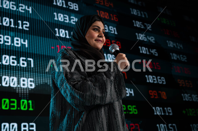 ندوات اقتصادية سعودية، سيدة اعمال خليجية سعودية عربية تمسك المايكروفون وتتحدث في مؤتمر اقتصادي، مقترحات ونصائح حول التدوال الإلكتروني، عرض تقديمي لأخبار أسهم الشركات في البورصة السعودية، خلفية شاشات الكترونية حديثة لمتابعة السوق السعودي