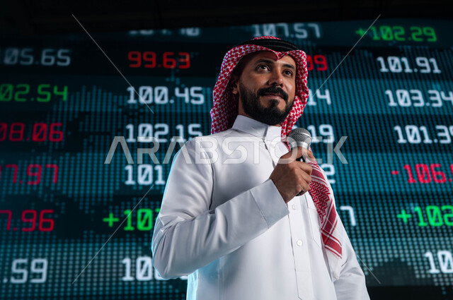 ندوات اقتصادية سعودية، مقترحات ونصائح حول التدوال الإلكتروني، رجل اعمال خليجي سعودي عربي يمسك المايكروفون ويتحدث في مؤتمر اقتصادي، خلفية شاشات الكترونية حديثة للأسهم المتداول بها في السعودية، عرض تقديمي لأخبار أسهم الشركات في البورصة السعودية