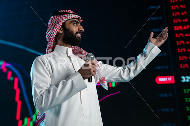 مقترحات ونصائح حول التدوال الإلكتروني، رجل اعمال خليجي سعودي عربي يمسك المايكروفون ويتحدث في مؤتمر اقتصادي سعودي، عرض تقديمي لأخبار أسهم الشركات في البورصة السعودية، خلفية شاشات الكترونية حديثة لمتابعة صعود وهبوط مؤشر البورصة