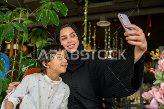 طفل عربي سعودي خليجي يقضي وقته مع امه في المطعم بايماءات و تعابير وجه مختلفة ، تلتلقط الام صور سلفي لها و لأبنها بكاميرا هاتفها المحمول، صورة تذكارية ، مطعم سعودي 
