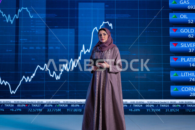 قراءة الموجز الأخباري بإستخدام التابلت، دمج البيئة العملية مع التقنية، مذيعة وإعلامية عربية خليجية سعودية تقوم بتقديم الأخبار الإقتصادية السعودية، التغيرات الحاصلة في أسعار السلع، خلفية شاشة إلكترونية حديثة لصعود وهبوط مؤشر الأسهم والبورصة