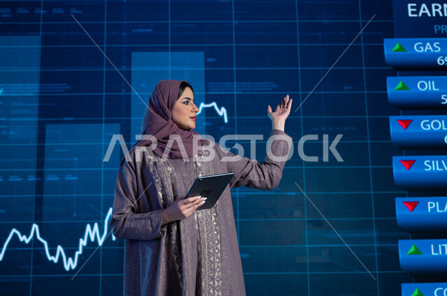 خلفية شاشة إلكترونية حديثة لصعود وهبوط مؤشر الأسهم والبورصة، مذيعة وإعلامية عربية خليجية سعودية تقوم بتقديم النشرات الإخبارية الإقتصادية، قراءة الموجز الأخباري بإستخدام جهاز التابلت، شرح وتوضيح التغيرات الحاصلة في أسعار السلع والثروات