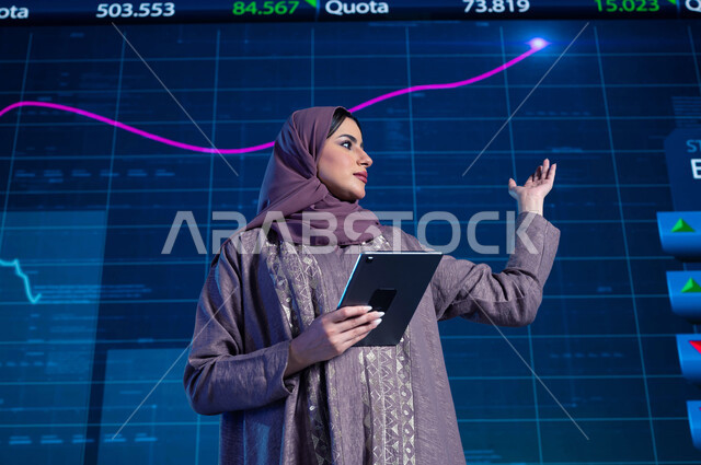 مذيعة وإعلامية عربية خليجية سعودية تقوم بتقديم النشرات الإخبارية الإقتصادية، قراءة الموجز الأخباري بإستخدام جهاز التابلت، شرح وتوضيح التغيرات الحاصلة في أسعار السلع والثروات، خلفية شاشة إلكترونية حديثة لصعود وهبوط مؤشر الأسهم والبورصة