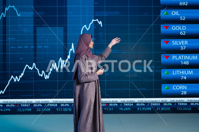مذيعة وإعلامية عربية خليجية سعودية تستخدم التابليت في قراءة الموجز الأخباري، التغيرات الحاصلة في أسعار السلع والثروات، تقديم الأخبار الإقتصادية السعودية، دمج البيئة العملية مع التقنية والتكنولوجيا، خلفية شاشة إلكترونية حديثة لصعود وهبوط مؤشر الأسهم