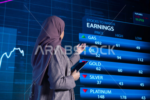 شرح وتوضيح التغيرات الحاصلة في أسعار السلع والثروات، مذيعة وإعلامية عربية خليجية سعودية تقوم بتقديم النشرات الإخبارية الإقتصادية، قراءة الموجز الأخباري بإستخدام جهاز التابلت، خلفية شاشة إلكترونية حديثة لصعود وهبوط مؤشر الأسهم والبورصة