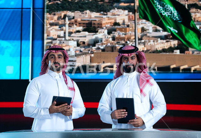Merging the practical environment with technology and technology, two Saudi Arabian Gulf broadcasters presenting the newsletter, reading the news brief using the tablet, celebrating the Saudi Flag Day and commemorating the National Day, an electronic scre