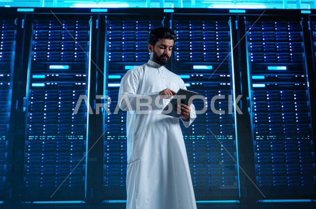 مفهوم  تكنولوجيا التخزين السحابي، رجل عربي خليجي سعودي يستخدم جهاز التابليت في الإشراف وإدارة معلومات الداتا سنتر، تسهيل الوصول للمعلومات من خلال نظم قواعد البيانات، خلفية هولوجرامية ثلاثية الأبعاد لمركز البيانات