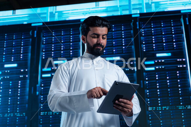 Benefiting from Saudi expertise in developing the data center, the cloud storage network, a Saudi Arabian Gulf man using a tablet, managing the servers control room and giant internet servers, a 3D hologram background for the data center