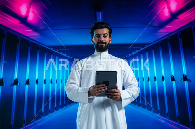 إستثمار الخبرات السعودية في بناء مراكز بيانات متطورة وحديثة، شبكة التخزين السحابي، خلفية هولوجرامية ثلاثية الأبعاد لغرفة التحكم بالسيرفرات وخوادم الانترنت، رجل عربي خليجي سعودي يستخدم جهاز التابليت في الإشراف وإدارة معلومات الداتا سنتر