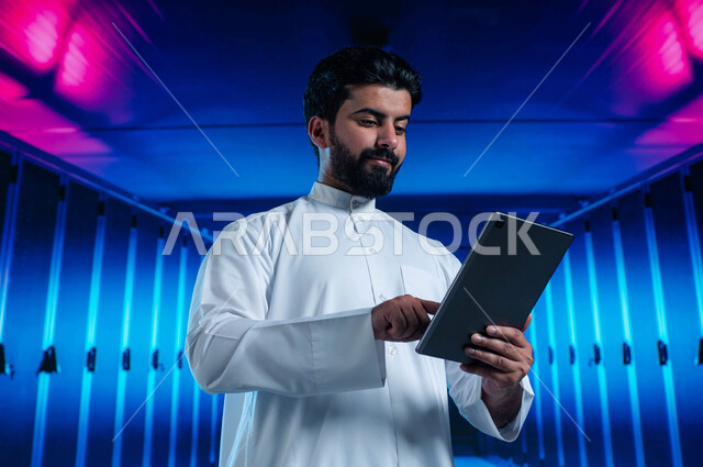 غرفة التحكم بالسيرفرات وخوادم الانترنت، تسهيل الوصول للمعلومات من خلال نظم قواعد البيانات، رجل عربي خليجي سعودي يقوم بالتحكم بالمواقع المستضافة على السيرفرات من خلال جهاز التابلت، خلفية هولوجرامية ثلاثية الأبعاد لمركز البيانات