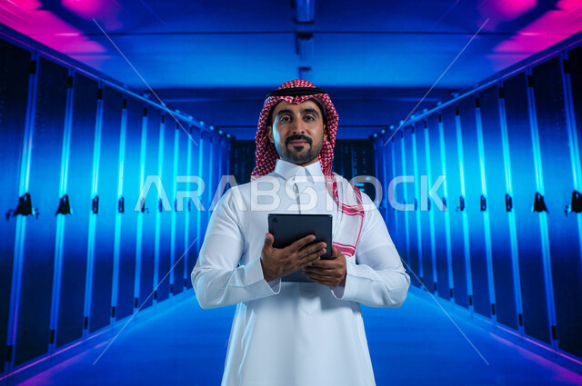خلفية هولوجرامية ثلاثية الأبعاد لغرفة الخوادم والسيرفرات، رجل عربي خليجي سعودي يقوم بالإشراف وإدارة بيانات الداتا سنتر باستخدام جهاز التابليت، مفهوم  تكنولوجيا التخزين السحابي، خوادم الانترنت العملاقة، حفظ وتخزين المعلومات لمستخدمين المواقع