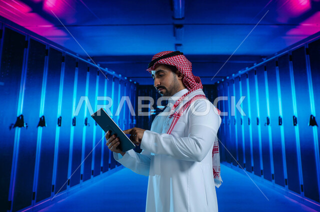 خوادم الانترنت العملاقة، رجل عربي خليجي سعودي يقوم بالإشراف وإدارة بيانات الداتا سنتر باستخدام جهاز التابليت، مفهوم  تكنولوجيا التخزين السحابي، حفظ وتخزين المعلومات لمستخدمين المواقع، خلفية هولوجرامية ثلاثية الأبعاد لغرفة الخوادم والسيرفرات
