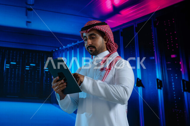 حفظ وتخزين المعلومات لمستخدمين المواقع، رجل عربي خليجي سعودي يقوم بالإشراف وإدارة بيانات الداتا سنتر باستخدام جهاز التابليت، مفهوم تكنولوجيا التخزين السحابي، خوادم الانترنت العملاقة، خلفية هولوجرامية ثلاثية الأبعاد لغرفة الخوادم والسيرفرات
