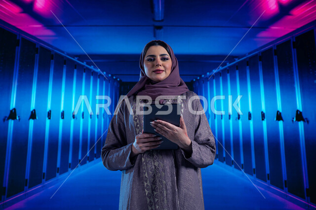 خوادم الانترنت العملاقة، امرأة عربية خليجية سعودية تقوم بالإشراف وإدارة بيانات الداتا سنتر باستخدام جهاز التابليت، مفهوم  تكنولوجيا التخزين السحابي، حفظ وتخزين المعلومات لمستخدمين المواقع، خلفية هولوجرامية ثلاثية الأبعاد لغرفة الخوادم والسيرفرات