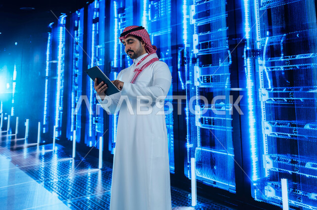 خلفية هولوجرامية ثلاثية الأبعاد لغرفة الخوادم والسيرفرات، تسهيل الوصول للمعلومات من خلال نظم قواعد البيانات، رجل عربي خليجي سعودي يقوم بالإشراف والتحكم في مركز البيانات باستخدام جهاز التابليت، الاستفادة من الخبرات السعودية في تطوير الداتا سنتر