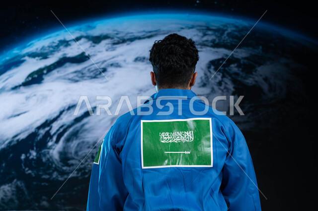 رحله السعودية الى الفضاء الخارجي, شعار علم السعودية على البدلة الفضائية الزرقاء, صورة من الخلف لرائد فضاء عربي خليجي سعودي في العالم الفضائي, تطور و نمو و دعم قطاع الفضاء في الوطن العربي, رواد فضاء سعوديين, خلفية هولوجرامية ثلاثية الابعاد لكوكب الارض.