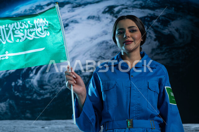 رحلة السعودية نحو العالم الخارجي, النظر الى الكاميرا, رائدة فضاء عربية خليجية سعودية ترتدي بدلة الفضاء السعودية تحمل بيدها علم المملكة العربية السعودية في الفضاء الخارجي, رواد فضاء سعوديين, خلفية هولوجرامية ثلاثية الابعاد لكوكب الارض