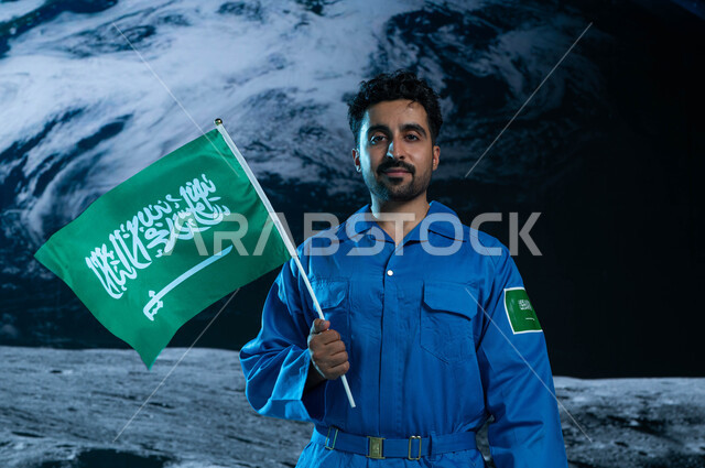 رحلة السعودية نحو العالم الخارجي, النظر الى الكاميرا, رائد فضاء عربي خليجي سعودي يرتدي بدلة الفضاء السعودية يحمل بيده علم المملكة العربية السعودية في الفضاء الخارجي, رواد فضاء سعوديين, خلفية هولوجرامية ثلاثية الابعاد لكوكب الارض