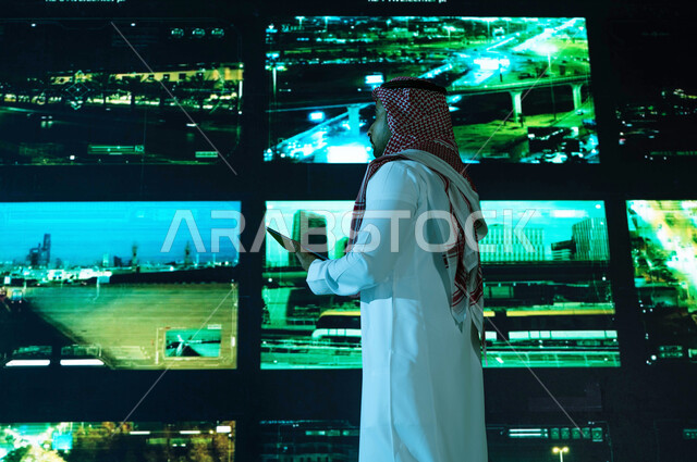 Carrying out traffic control in the roads and streets of the city of Riyadh, the background of modern screens, a picture from the back of a Saudi Arabian Gulf man wearing traditional dress, advanced electronic cameras, the center of administration and sup