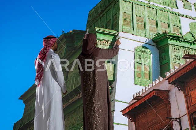 مناطق سياحية مشهورة, الاشارة الى شيء ما, التصميم المعماري العربي القديم, المعالم التاريخية في جدة في حي البلد, صورة من الخلف لزوجان عربيان خليجيان سعوديان يقومان بالنظر إلى المباني الأثرية في البلد التاريخي في مدينة جدة في المملكة العربية السعودية