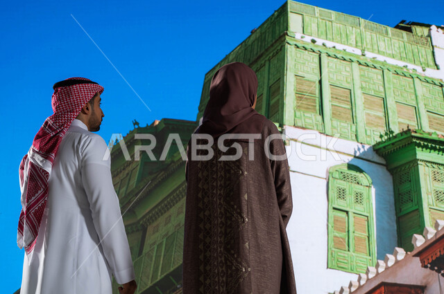 أماكن سياحية مشهورة, التصميم المعماري العربي القديم, المعالم التاريخية في جدة في حي البلد, صورة من الخلف لرجل و امرأة عربيان خليجيان سعوديان يقومان بالنظر إلى المباني الأثرية في البلد التاريخي في مدينة جدة في المملكة العربية السعودية