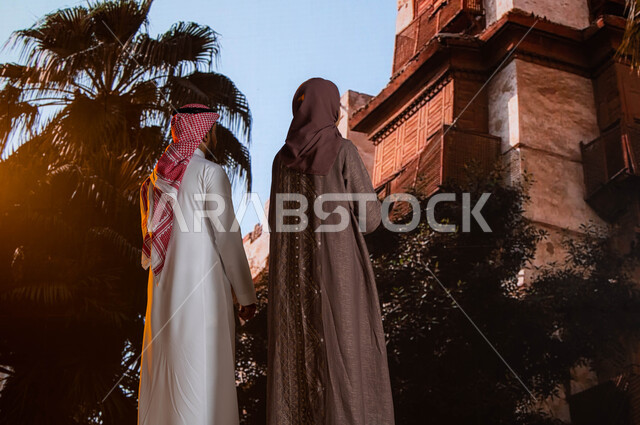 التشجير وزراعة النخيل, التصميم المعماري العربي القديم, مناطق سياحية مشهورة, صورة من الخلف لزوجان عربيان خليجيان يقفان أمام المعالم التاريخية في جدة في حي البلد، النظر إلى المباني الأثرية في البلد التاريخي في مدينة جدة