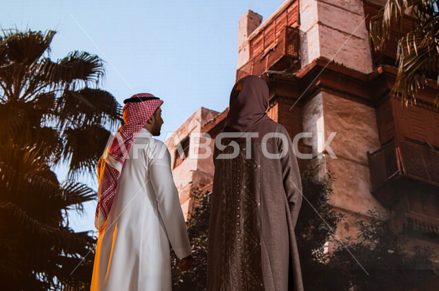 التصميم المعماري العربي القديم, التشجير وزراعة النخيل, مناطق سياحية مشهورة, صورة مقربة من الخلف لزوجان عربيان خليجيان يقفان أمام المعالم التاريخية في جدة في حي البلد، النظر إلى المباني الأثرية في البلد التاريخي في مدينة جدة