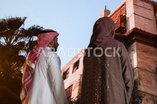 النظر إلى المباني الأثرية في البلد التاريخي في مدينة جدة, التصميم المعماري العربي القديم, التشجير وزراعة النخيل, مناطق سياحية مشهورة, صورة من الخلف لزوجان عربيان خليجيان يقفان أمام المعالم التاريخية في جدة في حي البلد