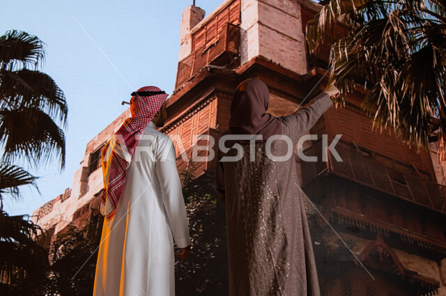 الاشارة الى شيء ما، التشجير وزراعة النخيل, التصميم المعماري العربي القديم, مناطق سياحية مشهورة, صورة من الخلف لزوجان عربيان خليجيان يقفان أمام  المعالم التاريخية في جدة في حي البلد، النظر إلى المباني الأثرية في البلد التاريخي في مدينة جدة