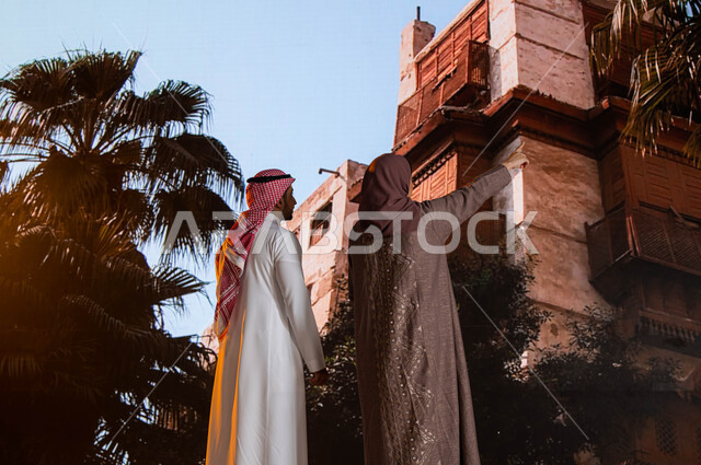 التشجير وزراعة النخيل, الاشارة الى شيء ما, التصميم المعماري العربي القديم, مناطق سياحية مشهورة, صورة من الخلف لزوجان عربيان خليجيان يقفان أمام  المعالم التاريخية في جدة في حي البلد، النظر إلى المباني الأثرية في البلد التاريخي في مدينة جدة