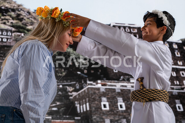 قدوم السياح من شتى أنحاء العالم، الوقوف أمام الأثار و البيوت التاريخية القديمة في أبها ،صورة مقربة لولد عربي خليجي يرتدي الزي الشعبي الجنوبي مع سائحة أجنبية في  قرية رجال ألمع التراثية في عسير،عادات وتقاليد شعبيه في ارتداء اكليل من الورد العسيري العطري