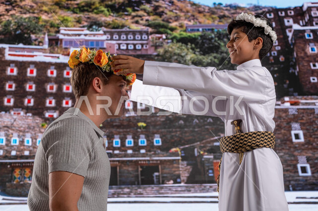 عادات وتقاليد شعبيه في ارتداء اكليل من الورد العسيري العطري ،الأثار و البيوت التاريخية القديمة في أبها ،ولد عربي خليجي يرتدي الزي الشعبي الجنوبي مع سائح أجنبي بإيماءات تدل على السعادة ,مناطق سياحية مشهورة, الوقوف أمام قرية رجال ألمع التراثية في منطقة عسير