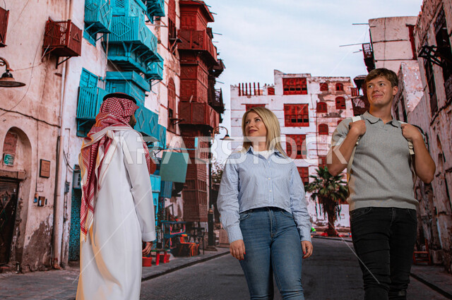 مناطق ومعالم سياحيه تاريخيه مشهوره في السعودية، صورة من الخلف لرجل سعودي مع السائحين الاجانب يتجولون بين الأحياء الشعبيه و المباني الأثرية في جده التاريخيه, الرواشين الخشبيه في البناء المعماري العربي القديم  في حي البلد, الدليل السياحي للسياح
