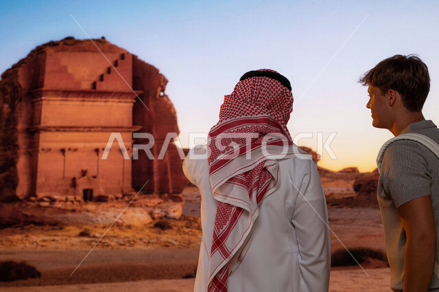 معالم قديمة أثرية تاريخية، الوقوف أمام قصر الفريد في محافظة العلا في السعودية ,صورة مقربة من الخلف لرجل عربي خليجي مع سائح اجنبي يقوم بزيارة الطبيعة الصحراوية في الأماكن السياحية في السعودية ويشير إلى شيء ما, الدليل السياحي للسياح، انشطه ترفيهيه ممتعه