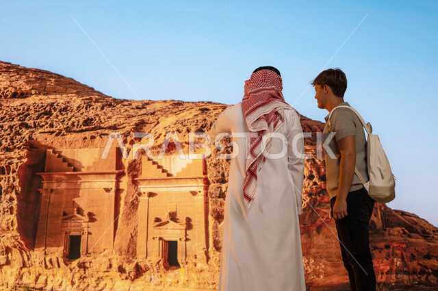 الأماكن القديمة الأثرية، المعالم التاريخيه الاسلامية البارزه في مدائن صالح في العلا، انشطه ترفيهيه سياحية في السعودية, صورة من الخلف لمرشد سياحي سعودي مع سائح أجنبي يقفان أمام الطبيعة الجبلية المميزة ويشير إلى شيء ما، فتح أبواب السياحة السعودية