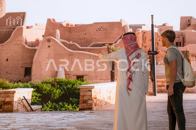 المباني الطينيه و الآثار في حي البجيري في مدينة الدرعية في الرياض، معلم و منتزه تراثي تاريخي على الطراز الشعبي القديم، مركز وطني ثقافي سياحي ،صورة من الخلف لزائر اجنبي مع رجل عربي خليجي سعودي يرتدي الزي التقليدي يقف امام قصر سلوى و الإشارة الى شيء ما