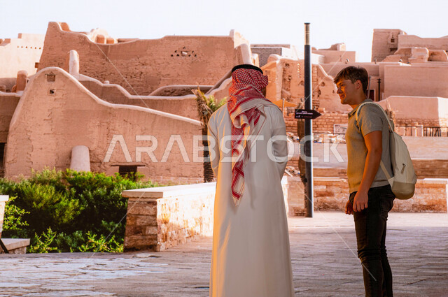 A historical heritage landmark and park in the old folk style, a national, cultural and tourist center. A picture from the back of a foreign visitor with a Saudi Arabian Gulf man wearing traditional dress standing in front of Salwa Palace exchanging conve