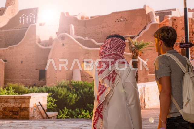 مركز وطني ثقافي سياحي ،صورة من الخلف لزائر اجنبي يرتدي حقيبة الظهر الشخصية مع رجل عربي خليجي سعودي يرتدي الزي التقليدي يقف امام قصر سلوى، الآثار و المباني الطينيه في حي البجيري في مدينة الدرعية في الرياض، منتزه ومعلم تاريخي تراثي على الطراز الشعبي القديم
