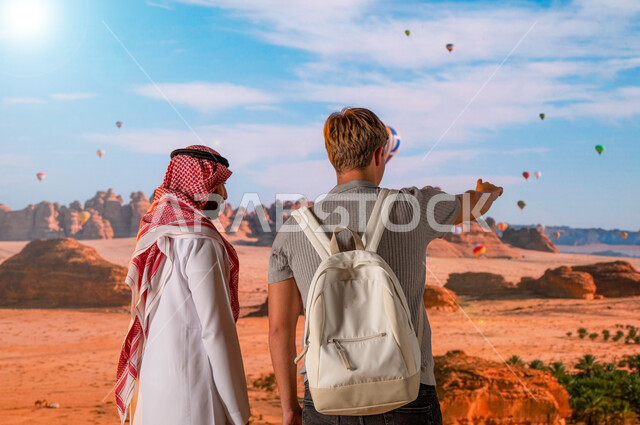 فعاليات و انشطه ترفيهيه، الطيران في بالون الهواء الساخن, الدليل السياحي للسياح، المناطيد في سماء المملكة في محافظة العلا, صورة من الخلف لرجل عربي خليجي يرتدي الزي السعودي التقليدي مع سائح أجنبي يشير إلى شيئ ما , معالم أثرية تاريخية قديمة .